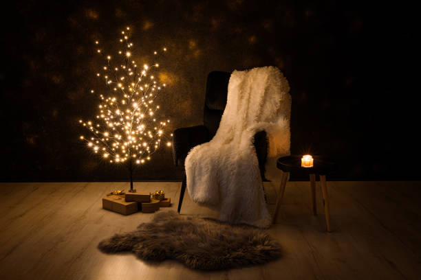 Alternative Christmas tree concept. Minimalist black color wire tree with lights illuminated at night. Black armchair, white plaid, presents under the tree, candle burning. Cozy Christmas Eve.