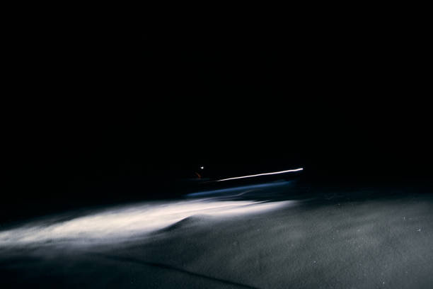 A car drives along a desolate road at night, its headlights cutting through the pitch darkness, revealing the empty asphalt and surrounding shadows.