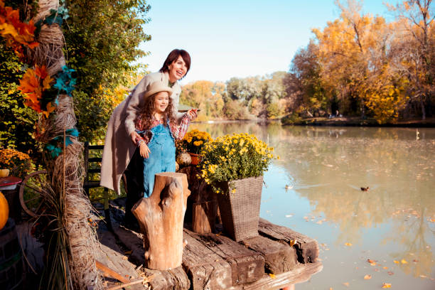 Mother and her girl enjoying lake view in Autumn day. Standing on the pier decorated with pumpkins and fall flowers