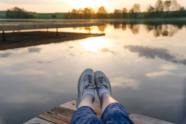 close-up view of legs in white sneakers, young beautiful caucasian woman sits at wooden pier on river bank in summer at sunset, green trees in background, summer holidays, travelling concept