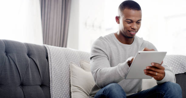istock House, typing and black man on sofa, tablet and connection with website info, relax and social media in lounge. African person, apartment and guy on couch, technology and communication with network 2184536078