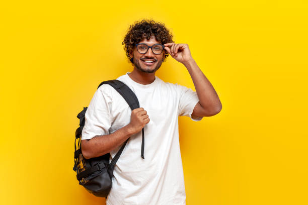 istock indian guy student in a white t-shirt with glasses and with backpack points back on yellow isolated background, curly man goes to college to study 2184538273