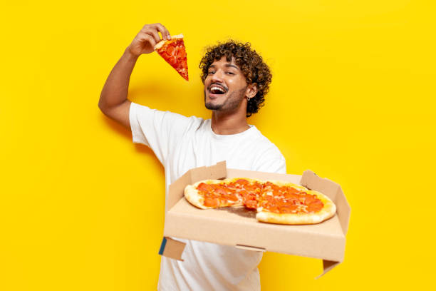 istock indian man eating delicious pizza on yellow isolated background, curly guy student holding pizza box and biting a piece of fast food 2184538367