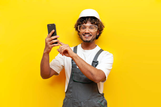 istock Indian male builder in hard hat and overalls using smartphone on yellow isolated background, Indian craftsman in uniform selects online on mobile 2184538545