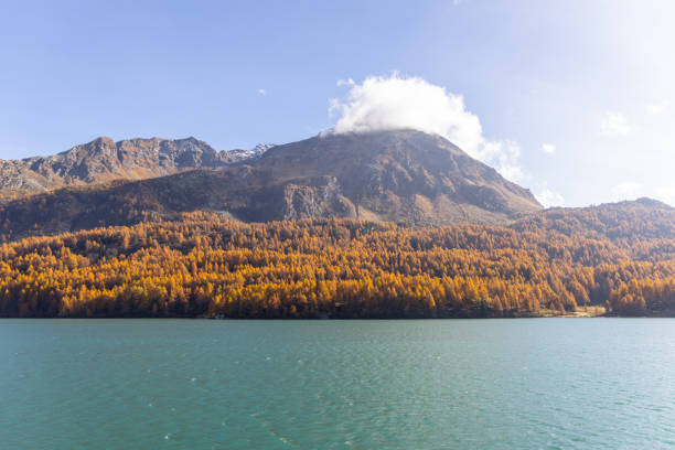 Serene autumn landscape featuring a calm lake with a backdrop of vibrant orange and yellow trees, a rugged mountain, and a cloud-covered peak. A peaceful scene of nature's seasonal beauty.