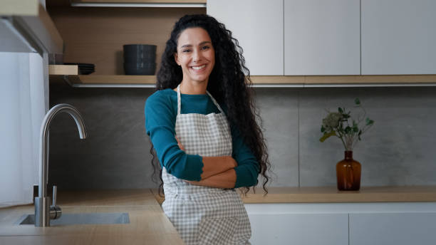 Smiling young woman maid housewife mistress baker housekeeper with long curly hair wearing checkered apron standing with folded arms confident chef posing in modern domestic kitchen cuisine interior. High quality 4k footage mujer joven sonriente, ama de casa, ama de casa, amante, panadera, ama de llaves, con cabello largo y rizado, con delantal a cuadros, de pie con los brazos cruzados, chef seguro posando en el interior de la cocina de la cocina doméstica moderna