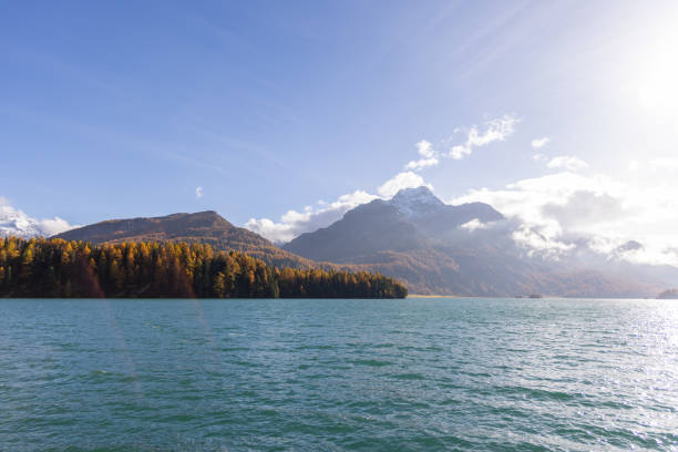 The sun shines brightly over a serene mountain lake, surrounded by autumn-colored forests and snow-tipped peaks under a clear sky.