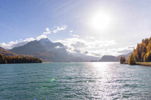 The sun shines brightly over a serene mountain lake, surrounded by autumn-colored forests and snow-tipped peaks under a clear sky.
