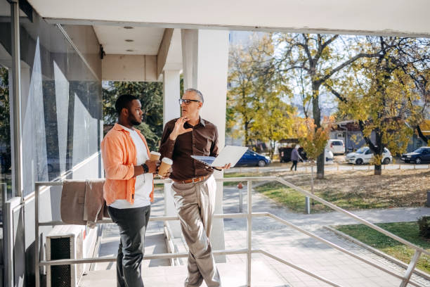 istock Older and younger male colleagues working together and sharing experience as well as  providing mentorship to younger colleague while standing on coffee break in front of company building 2184564457