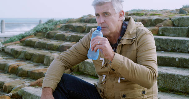 istock Senior, man and drinking with water on stairs by beach for hydration, thirst or natural sustainability. Mature, male person or relax with mineral liquid, beverage or refreshment on steps by ocean 2184576255