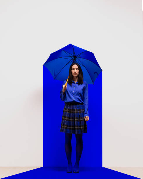istock Woman dressed in blue standing under blue umbrella in blue square spotlight. Introspection, isolation, and resilience in minimalist setting. Contemporary art collage. 2184580552