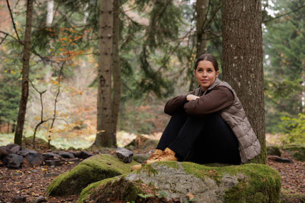 A woman enjoys a peaceful moment sitting on a mossy rock in a tranquil forest, surrounded by tall trees. The scene captures the serenity of nature and the beauty of solitude.