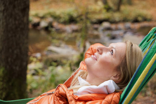 A person relaxes in a hammock surrounded by serene autumn woods, next to a gentle stream. The scene exudes peace and nature's soothing charm, with a backpack to the side, suggesting a day of adventure