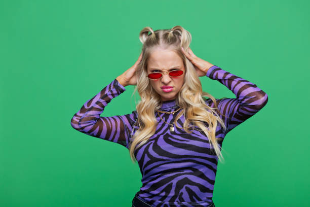 Portrait of disappointed young blonde woman wearing purple animal pattern blouse and red sunglasses looking at camera with raised hands. Studio shot, green background.