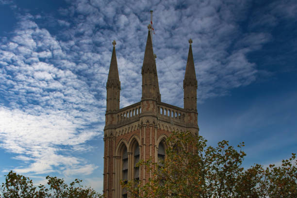A low angle view of a stone church tower with three spires and a flag on top, against a bright blue sky with white, puffy clouds. Tree branches are visible in the foreground.