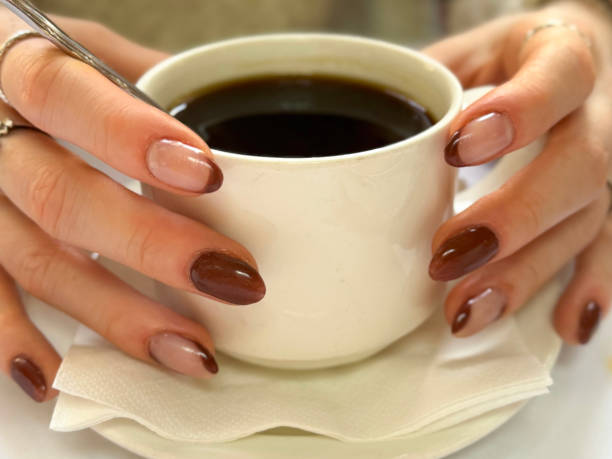 istock Full frame image of unrecognisable woman's hands holding white cup and saucer with napkin, black Americano coffee with stainless steel teaspoon, showing nail varnish manicure design, cafe setting, focus on foreground 2184616407