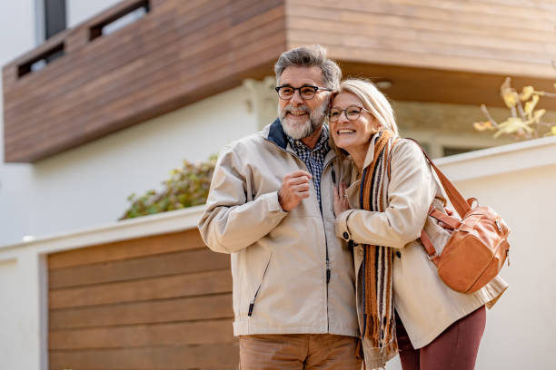Senior couple embracing and smiling in front of a modern house on a sunny day, showcasing warmth, love, and happiness in a relaxed outdoor setting. stock photo