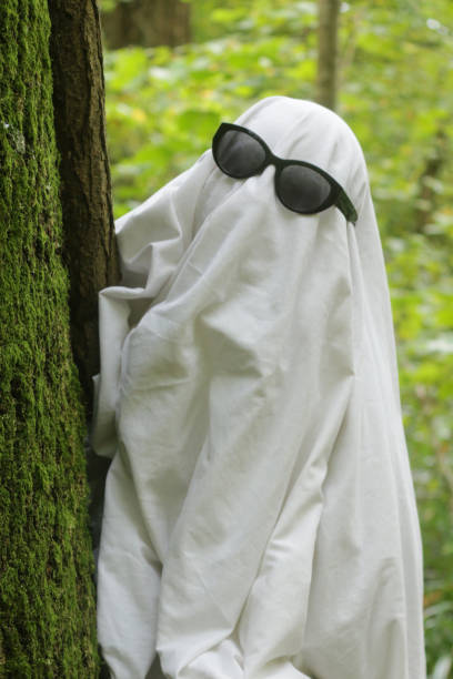 Stock photo showing close-up view of a Halloween woodland scene with an unrecognisable person dressed as a ghost by being covered by a white bed sheet, with sunglasses for the eyes. This ghoul is pictured in a 'haunted forest' setting in the autumn, with freshly fallen brown, orange and yellow oak leaves covering the floor.