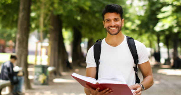 istock Smiling university student walking in a park reading a book 2184635068