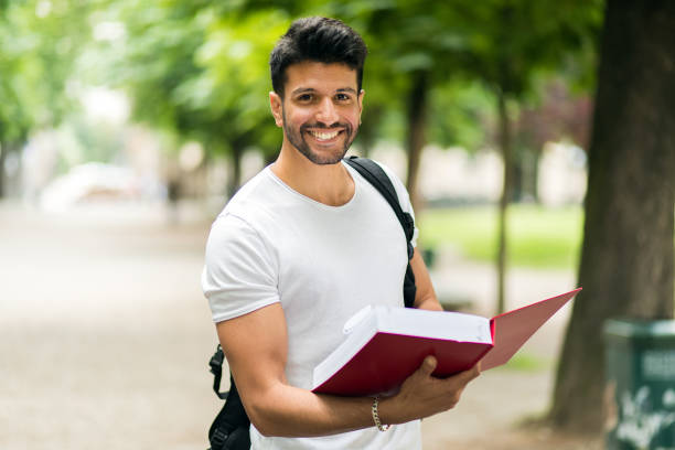 istock Smiling student holding a book and wearing a backpack in a park 2184637030