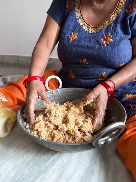 Image of unrecognisable Indian woman sat on floor wearing Salwar Kameez traditional clothing, making Indian sweets, ladoo recipe gram flour (besan), ghee and sugar mixture in karahi (Indian wok), focus on foreground stock photo