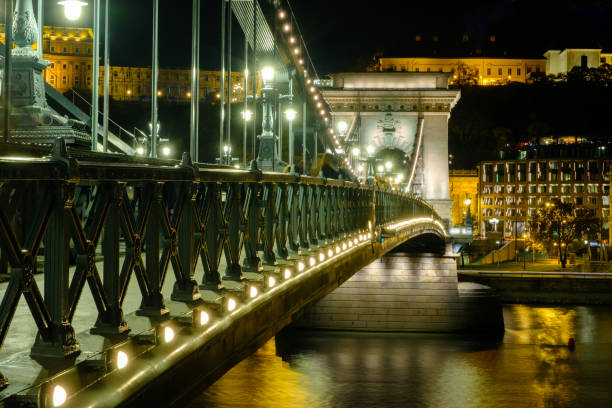 The Chain Bridge stands majestically over the Danube River, illuminated by streetlights at night. This iconic landmark connects Buda and Pest, showcasing stunning architecture and vibrant city life along the riverbank.
