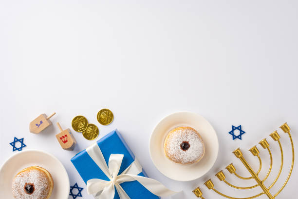 A festive Hanukkah setup featuring donuts, dreidels, chocolate coins, a menorah, and a gift box arranged on a white surface