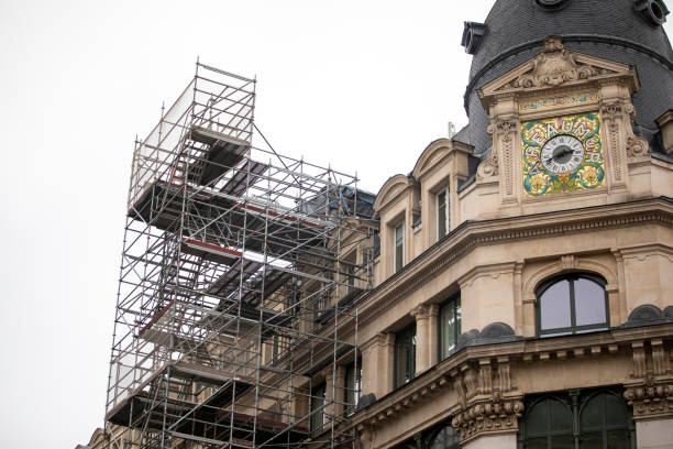 Budapest, Hungary - April 2025 A detailed view of a beige stone historic building facade featuring intricate architectural details such as carved moldings, arched windows, and wrought-iron balconies, surrounded by metal scaffolding for renovation work. A red fabric hangs from one balcony, adding a pop of color against the neutral tones of the stone and gray scaffolding. The scene captures an urban construction site on a narrow city street, with adjacent buildings visible in the background. Outdoors in a European urban setting, likely daytime. This image suits commercial uses in media coverage of city preservation projects, architecture features, urban development stories, or illustrations for books on historic buildings and renovation processes.