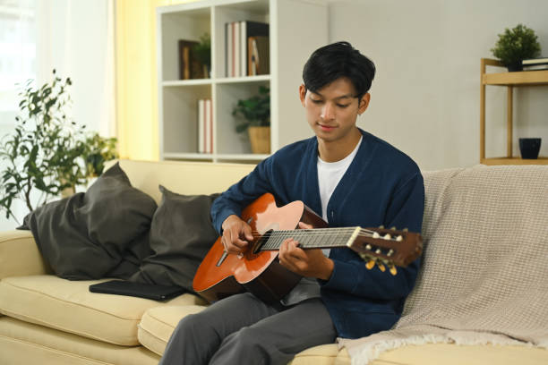 istock Young man wearing a blue sweater playing the guitar on a couch 2184696489