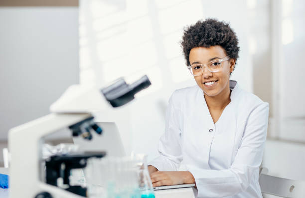 A smiling young woman with mixed ethnicity works diligently in a lab, clad in a white coat, with a microscope and scientific instruments nearby.