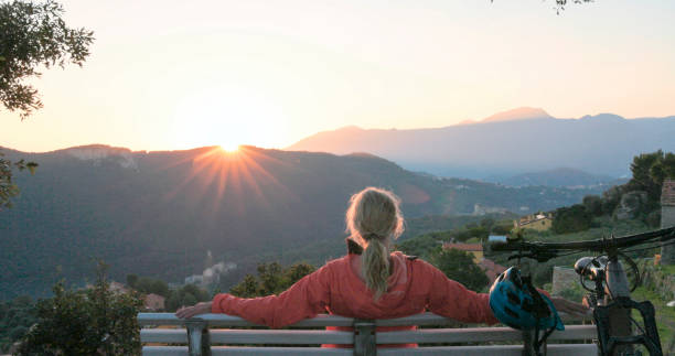 Mature woman mountain biker watches sunrise over distant hills from bench, Liguria