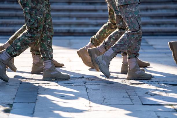Military personnel in camouflage uniforms marching in unison on a sunny day, casting shadows on the pavement.