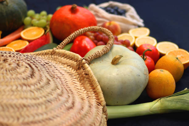 Round straw bag and various seasonal fruits and vegetables on dark background. Summer and fall produce. Selective focus.