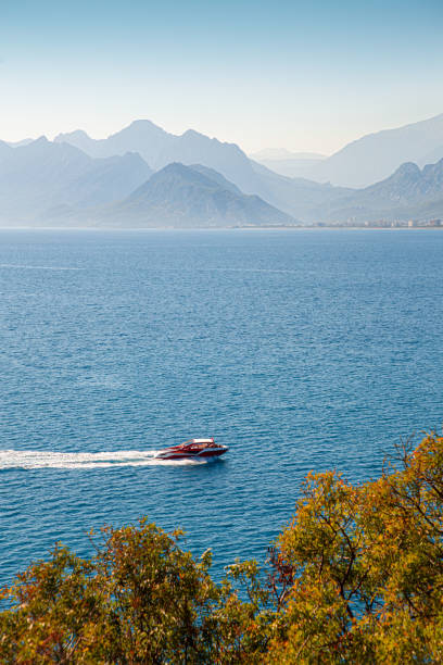A dynamic aerial view of a speedboat cutting through the crystal-clear waters of the Mediterranean Sea, with rugged rock shores stretching into the distance. The bright blue sea contrasts with the dramatic cliffs, creating a breathtaking natural backdrop. The speedboat’s motion adds energy to the scene, emphasizing the adventurous side of Antalya’s coastal beauty and offering a thrilling contrast to the serene surroundings.Ideal for promoting adventure tourism, water sports, luxury vacations, speedboat rentals, and showcasing the exciting side of Turkey’s Mediterranean coastline.