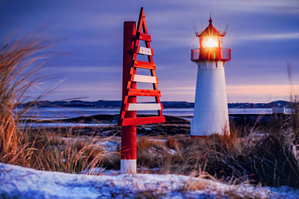 Coastal lighthouse glowing warmly against winter twilight, with striking red and white triangular navigational marker beside it, set amidst snow-dusted dunes, frosty grasses and purple skies.Sylt.