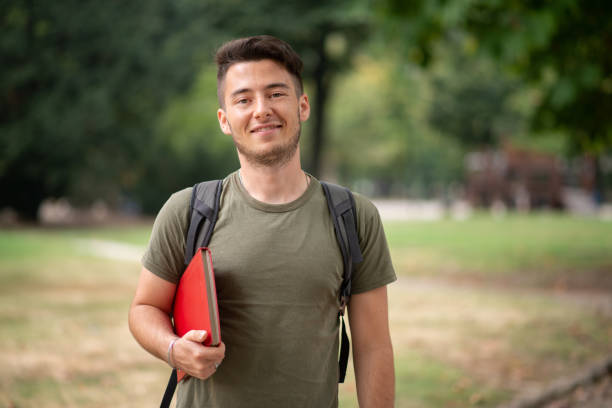 istock Smiling university student holding book and wearing backpack in park 2184725950