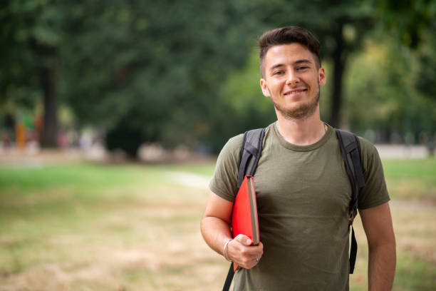 Smiling university student walking in park with backpack and book