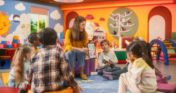 istock Happy Professional Kindergarten Teacher Using Tablet Computer to Show Numbers and Alphabet Characters to Smart Little Kids at a Modern Colorful Kindergarten. Female Educator Developing Social Skills 2184737517
