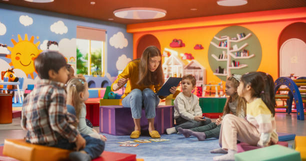 istock Beautiful Daycare Teacher Using a Tablet Computer to Tell an Educating Story for Young Talented Kids. Female Educator Having Fun Supervising Diverse Group of Small Boy and Girls 2184737567