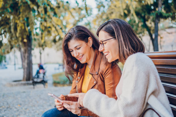 istock Two cheerful woman talking and using phone while resting in the park 2184745151