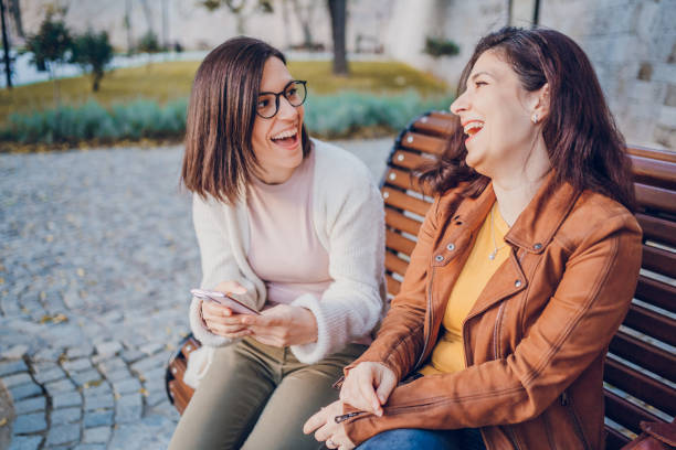 istock Two cheerful woman talking and using phone while resting in the park 2184745172
