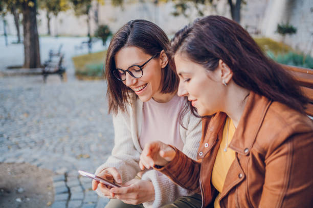 istock Two cheerful woman talking and using phone while resting in the park 2184745246