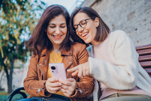 istock Two cheerful woman talking and using phone while resting in the park 2184745329