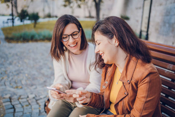 istock Two cheerful woman talking and using phone while resting in the park 2184745415