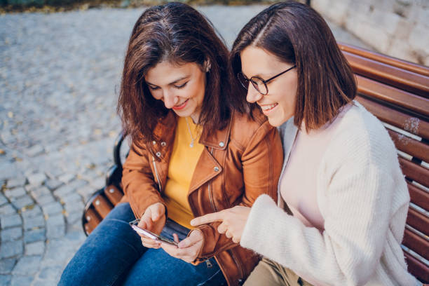 istock Two cheerful woman talking and using phone while resting in the park 2184745446