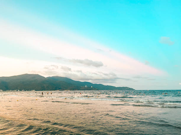The serene morning paints the sky with vibrant shades of orange, pink, and purple, reflecting on the calm waters of My Khe Beach. Silhouettes of swimmers and fishing boats dot the tranquil coastline.