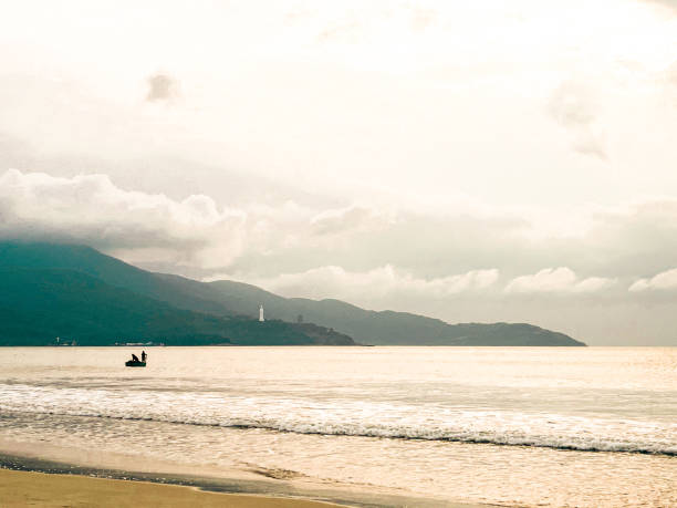 The serene morning paints the sky with vibrant shades of orange, pink, and purple, reflecting on the calm waters of My Khe Beach. Silhouettes of swimmers and fishing boats dot the tranquil coastline.
