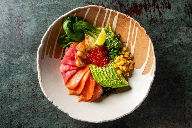 A bowl of poke with raw fish slices, avocado, corn, greens, and rice, garnished with sesame seeds. The vibrant colors and fresh ingredients make it a visually appealing and healthy meal option.