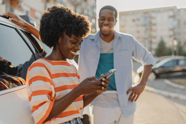 Woman typing on phone with man leaning on car