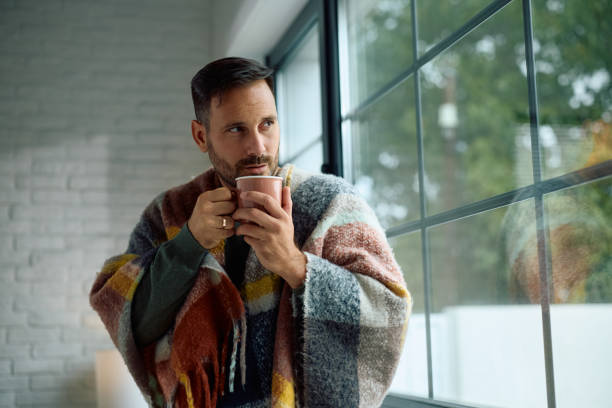 Pensive man wrapped in a blanket drinking tea by the window.